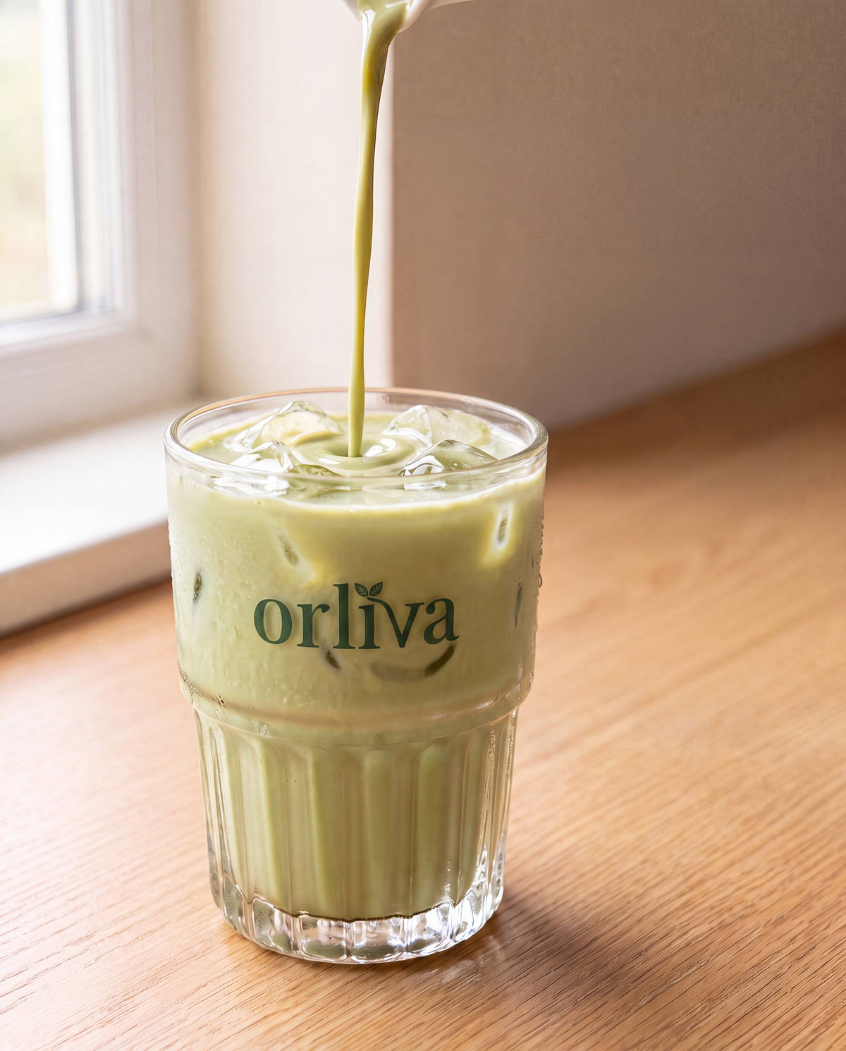 Iced ORLIVA matcha latte in a ribbed glass, creamy milk swirl, ORLIVA pouch softly blurred behind